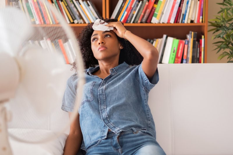 Young woman reclined in front of fan with towel on her forehead.