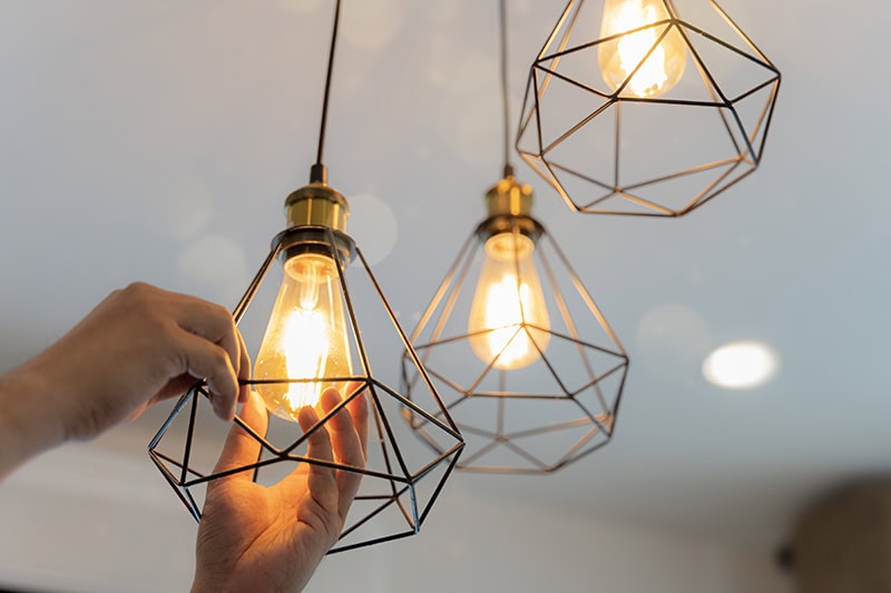 A hand adjusting or replacing a glowing Edison-style bulb inside a modern geometric wire-cage pendant light fixture, surrounded by similar lit hanging lights.